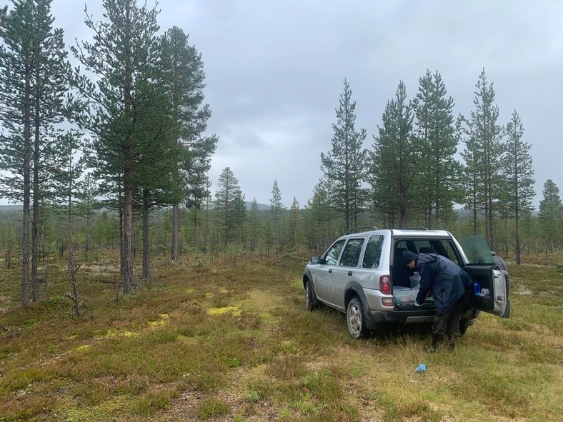 En silvrig bil står i skogsbrynet. En man plockar ut material till sitt forskningsprojekt ut bakluckan.