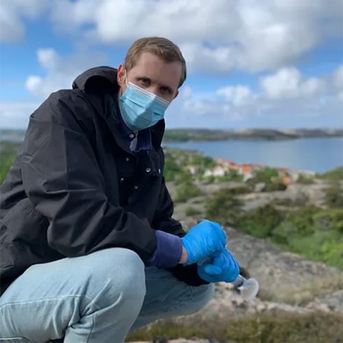 Patrick på en klippa i skärgården, ute på ett eDNA projekt. Han bär ett blått munskydd och blåa handskar.